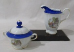 PORCELANA - REAL S. PAULO - Um açucareiro e uma leiteira em porcelana nacional vitrificada branca, apresentando cena galante em rica policromia ao centro, seguida de friso dourado, bordas ligeiramente onduladas e adornadas com larga faixa na tonalidade azul, finalizando com filetes em pintura à ouro, pega da tampa em formato de flor, alças trabalhadas em folhas com vasta douração, marcados no fundo. Med. leiteira: alt total 15 cm x larg total 16 cm/ açucareiro: alt total 11,5 cm x larg total 15 cm.