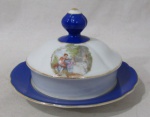 PORCELANA - REAL S. PAULO - Uma manteigueira em porcelana nacional vitrificada branca, apresentando cena galante em rica policromia ao centro, seguida de friso dourado, bordas ligeiramente onduladas e adornadas com larga faixa na tonalidade azul, finalizando com filetes em pintura à ouro, pega da tampa em formato de flor, marcada no fundo. Med. alt total 9 cm x diam total 15 cm.