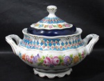 PORCELANA - Elegante terrina em porcelana vitrificada branca, apresenta roca decoração de motivos vegetalistas e geométricos em exuberante policromia e realces em douração, barrados adornados por largas faixas na tonalidade azul cobalto, arrematada com generosos filetes e frisos em pintura à ouro, bordas e base com movimento ondular, pega da tampa em forma estilizada de botão de flor. Medidas: alt total 21 cm x larg total 30 cm.