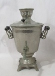 METAL PRATEADO - Um (1) Samovar russo decorativo em metal prateado, apoiado sobre base quadrada e pés em garras, com alça fixa. Falta torneira  Marcação no metal no centro. Altura 36 cm.