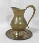BRONZE - Uma jarra em bronze contendo alça lateral estilizada e vazada, acompanha prato. Medidas da jarra. Altura: 23 cm: comprimento: 19 cm: medida do prato: diâmetro: 23 cm.