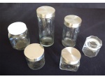 Seis potes para mantimentos de vidro sendo 4 com tampa inox, 1 com tampa de plástico branca e 1 tampa de vidro com bordas oitavadas. Alt. 27 a 11 cm.