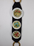 Três pratos decorativos em porcelana branca policromada decorada com rosas fixados sobre faixa de tecido vertical. Medida da faixa 93 x 17 cm e diam. do maior prato 19 cm.