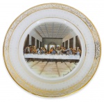 PORCELANA REAL - Elegante prato para decoração em porcelana branca esmaltada ricamente adornada com reprinte de A Santa Ceia de Leonardo Da Vinci e contorno em vibrante ouro. Possui registro da manufatura na base. Mede 23cm de diâmetro.