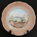 Gracioso prato para parede eou coleção em porcelana esmaltada em rosê, adornado à mão com paisagem com figura humana, com borda em movimento. Possui inscrição "Associação Comunitária Dos Moradores Do Bairro Mauá --- S. C. S. Mede 20 cm de diâmetro.