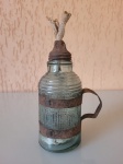 Antiga lamparina centenária, feita em vidro de água oxigenada da época com alça e bico em lata. Altura: 13,5cm; Circunferência: 17,5cm.