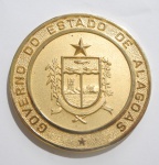 Moeda em bronze GOVERNO DO ESTADO DE ALAGOAS, no verso inscrições: ALAGOAS, TERRA DA PRODUÇÃO. Acondicionada em estojo. Med.: 5,5cm.