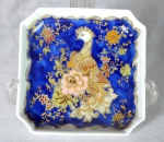Petisqueira alemã - KAISER em porcelana com pintura floral sobre-fundo azul borrão com realces em ouro. Chancela do fabricante no verso e ave fantasiosa ao centro. Med.: 3x14x14cm.