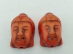 Duas peças em material sintético representando cabeças de Buda, com furação para pingente. Med. 3 x 2 cm.