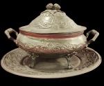 Imponente e antiga sopeira, ao estilo Português com prato (presentoir), confeccionada em Estanho, Corpo ovalado com rica decoração em relevo, apresentando motivos florais, arabescos e volutas, alças laterais trabalhadas e a terrina apoiada sobre quatro pés ornamentados. Medida maior: 43cm x 34cm.