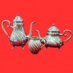 Antigo conjunto de chá e café em metal prateado apresenta um design clássico com corpos de formato bojudo e torção em caneluras, apoiados sobre pequenos pés, sendo um bule, um açucareiro e uma leiteira. Em metal espessurado a prata.