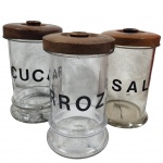 Conjunto contendo três potes antigos de mantimentos em vidro grosso, identificados como "AÇÚCAR", "ARROZ" e "SAL", acompanha suas respectivas tampas vedante, entalhadas em madeira. Medida: 29cm x 18cm. Obs, apresenta fio de cabelo.