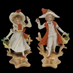 Par de delicados bibelôs, representando casal com trajes de época, sendo dama com Veste de saia ampla com corpete estruturado e chapéu adornado, refletindo a moda da corte francesa, onde o vestuário era uma linguagem de status e poder, cavalheiro Traja um casaco longo sobre calções e colete, conjunto clássico da indumentária masculina nobre do século XVIII. Peças confeccionadas em porcelana, em rica policromia feita a mão Medida: 12cm.