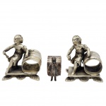Conjunto de três peças antigas em metal prateado, composto por dois porta-guardanapos figurativos da era vitoriana e um anel com figura feminina. O par apresenta o motivo detalhado de um Jovem Marinheiro, enquanto a terceira peça traz a imagem delicada de uma menina em pé, vestida com traje típico do século XIX e acoplada à frente do anel. Todas as peças foram produzidas pela renomada manufatura norte-americana Reed & Barton, fundada em 1824 em Taunton, Massachusetts, reconhecida mundialmente pela excelência em ourivesaria e objetos de mesa. Medidas: 10 cm x 10 cm.