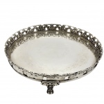 Antiga bandeja de servir Licor, circular em metal prateado, com borda vazada, trabalhada em intrincado padrão de galeria que remete a volutas e formas orgânicas. No centro, destaca-se uma delicada gravação floral em semicírculo, sustentada por pés decorativos robustos, com design clássico em relevos acentuados que sugerem influência dos estilos barroco. Medida: 35cm x 6cm.