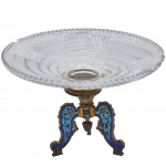 Centro de mesa em cristal lapidado, com base ornamentada em bronze e acabamento cloisonné em esmalte policromado. Apresenta rica decoração em tons de azul, turquesa e rosa, formando motivos florais e arabescos. Estrutura composta por três pés curvos. Medidas: 17 cm x 30 cm. obs. centro apresenta bicado.