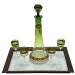 CRISTAL - Conjunto de licor composto por licoreira e quatro taças, decorado com frisos. Acompanha bandeja em bronze com alças ornamentadas. Medidas: bandeja 20 cm x 30 cm; licoreira 27 cm. Observação: uma taça apresenta bicado imperceptível.