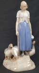 BING & GRONDAHL (B & G) - LINDA ESCULTURA EM PORCELANA REPRESENTANDO  CAMPONESA AFASTANDO GANSOS COM UMA VARA. MARCAS DA MANUFATURA SOB A BASE E ETIQUETA DA ANTIGA JOALHERIA CASTRO.  DINAMARCA, MEADOS DO SEC. XX. 24 CM DE ALTURA. UM MÍNIMO BICADO DEBAIXO DA CAUDA DE UM DOS GANSOS IMPERCEPTIVEL.