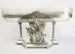 PALACIANO CENTRO DE MESA DE MUITO GRANDE DIMENSÃO EM METAL ESPESSURADO A PRATA COM MARCAS DA MANUFATURA WMF. CRISTAL ORIGINAL COM LAPIDAÇÃO FORMANDO LAURÉU A TODA VOLTA. BASE TEM DOIS CAVALEIROS MEDIEVAIS SOBRE CAVALOS ENVERGANDO ARMADURA, ESCUDO E LANÇA PARA JUSTA. SUSTENTADA FOR COLUNAS EM TORCEIL. ALEMANHA, SEC. XIX. 61 X 34 CM