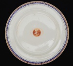 SERVIÇO DE DOM JOÃO VI - VISTA PEQUENA - PRATO EM PORCELANA CIA DAS ÍNDIAS QUE PERTENCEU AO SERVIÇO REAL DE D. JOÃO VI, CONHECIDO COMO VISTA PEQUENA. PERÍODO, QING, JIAQING (1796-1820). ABA COM BARRADO GEOMÉTRICO EM AZUL REALÇADO EM OURO, SUCEDIDO NA BORDA POR FRISO EM OURO. EM DIREÇÃO A CALDEIRA NOVO BARRADO FORMADO POR ELOS EM OURO ENTREMEADOS POR LOSANGO PONTILHADO EM AZUL. NA CALDEIRA DECORAÇÃO EM ELIPSE CONSTRUÍDA EM OURO E EM RESERVA, CENA NA TONALIDADE ROUGE DE FEUR COM CASA E ÁRVORES. REPRODUZIDO NO LIVRO LOUÇA DA ARISTOCRACIA DO BRASIL POR JENNY DREYFUS. CHINA, SEC. XIX, 25 CM DE DIAMETRO NOTA: De acordo com Leila Mezan Algranti, em seu artigo: Em torno da mesa do rei: artefatos, convivialidade e celebração no Rio de Janeiro joanino, em 26 de abril de 1821, após doze anos de estadia no Rio de Janeiro, iniciava-se a viagem de regresso de D. João VI a Portugal a fim de, mais uma vez, preservar sua Coroa e domínios. Para a autora, a partida da Corte foi precedida de muitas indecisões políticas e de intensos preparativos de ordem prática, da mesma forma como acontecera ao viajar para o Brasil, em 1807. Naquela ocasião, em meio aos riscos de invasão do Exército napoleônico, foi necessário acomodar nas embarcações não só os ilustres passageiros, mas também um conjunto imenso e variado de coisas destinadas ao uso particular de seus proprietários, bem como da Casa Real.Um conjunto de peças de prata, roupas de mesa e artigos de copa, recém-chegados do Rio de Janeiro, viajou para Lisboa sob a incumbência de Joze de Britto, fiel da mantearia do rei, e foi entregue no pátio das cozinhas do Palácio da Ajuda a Joze Caetano Trigo, também funcionário do mesmo departamento da Casa Real, o qual realizou o registro das peças e o assinou por ordem de João Lourenço de Andrade que, por sua vez, recebeu todo o material em nome de Caetano Joze de Campos e Andrade Pinto, manteeiro da Casa Real, naquele momento. O título do registro Inventário da Prata, Roupa e mais trem pertencente à Mantearia de Sua Majestade que veio do Ryo de Janeiro indica claramente que, ao retornar do Rio de Janeiro, D. João VI fez embarcar objetos de prata, os quais constituíam parte ou a totalidade de um serviço de mesa. Entretando deixou no país quase todos os serviços de porcelana adquiridos para seu uso aqui ou trasladados de Portugal quando de sua fuga. O conjunto poderia conter elementos da famosa baixela Germain, assim denominada por ter sido encomendada a François Germain por D. José I, em 1756, após o terremoto ter destruído a baixela de D. João V, fabricada pelo ourives de Luís XIV (Thomas Germain) e pai do prateiro de D. José. A suposição de que poderia haver peças da baixela Germain de D. José I entre os artefatos registrados no inventário advém do fato de que há informações sobre seu transporte para o Brasil, bem como estudos referentes à divisão da mesma entre D. João VI e D. Pedro I, antes do retorno do monarca a Portugal. Os bens que permaneceram no Brasil, por sua vez, teriam sido gravados com as insígnias imperiais e o monograma P1º. De acordo com Leonor d'Orey, considerando-se o que atualmente se encontra preservado desta baixela em Portugal, o lote do imperador do Brasil era menor, embora incluísse várias peças muito prestigiosas, as quais se encontram dispersas em coleções particulares e de museus estrangeiros, vendidas após a deposição de D. Pedro II. Sabe-se igualmente que entre a prataria de mesa de grande aparato pertencente à Casa Real havia peças da baixela dos duques de Aveiro, confiscada pela Coroa, após o atentado a D. José em 1759,15 assim como peças denominadas avulsas. Por outro lado, as baixelas para D. Pedro I foram encomendadas às famosas casas francesas, embora a técnica dos ourives radicados no Brasil fosse excelente, como apontou Francisco Marques dos Santos. Segundo o mesmo autor, muitas dessas peças emigraram, mas algumas voltaram. Havia, portanto, na corte no Rio de Janeiro, mais de um serviço de mesa em prata, sendo que um conjunto significativo dessas peças atravessou o Atlântico pelo menos duas vezes, a exemplo do que se sucedeu com a Família Real. Além disso, vários desses objetos foram possivelmente fabricados na França e na Inglaterra, uma vez que os prateiros portugueses nem sempre eram considerados, na época, suficientemente habilidosos para agradar o sofisticado gosto da alta nobreza portuguesa, consumidora de produtos de luxo importados não só de outros países da Europa, como do Oriente. Embora, como apontou Gonçalo de Vasconcelos e Sousa, houvesse prateiros de renome em Portugal, fabricando artefatos de mesa e mesmo baixelas completas, cuja qualidade do trabalho atingiu durante o século das Luzes um dos seus momentos mais importantes. De qualquer modo, a mesa da Casa Real e aquelas de outros membros das elites europeias e luso-brasileiras eram há muito tempo aparamentadas com artefatos de prata estrangeiros, bem como com porcelanas e vidros chineses, germânicos e franceses. Como bem apontou Nuno Vassallo e Silva, em seu estudo sobre a ourivesaria da mantearia da Casa de Aveiro, não é possível refletir sobre as baixelas de prata ou de porcelana usadas na mesa real sem levar em conta a conexão dessa indústria com as manufaturas europeias e orientais. Assim, é possível dizer que os objetos de luxo destinados ao serviço de mesa da Corte joanina, no Rio de Janeiro, resultavam de um circuito comercial e de comunicação que interligava diferentes impérios coloniais (português, inglês, francês), os quais se estendiam por três continentes: Europa, Ásia e América. No caso dos artefatos de prata que nos interessam comentar neste estudo, é provável que a matéria-prima utilizada fosse originária da América espanhola, tendo retornado ao mesmo continente após ter sido trabalhada por habilidosos artesãos franceses para uso e ostentação dos monarcas portugueses. Por outro lado, tais objetos indicam um movimento mais amplo de evolução do gosto e da maneira de viver dos membros da aristocracia europeia, que remonta ao final do século XVII, cuja etiqueta de mesa foi fixada na corte de Luís XIV. Como assinalou Marco Daniel Duarte, a alta sociedade quando se sentava à mesa se quer aprisionar por regras de etiqueta rígida. Mesmo levando-se em conta que no final do século XVIII Portugal acompanhava o restante da Europa, adotando nos banquetes reais a porcelana das índias e francesa como alternativas às baixelas de prata, conforme esclareceu Cristina Neiva, a prataria de mesa continuava a representar a riqueza e o poder das famílias reais, haja vista não só a encomenda de D. José I a François Germain, cuja baixela foi produzida ao longo da segunda metade do século XVIII, como também o fato de esta ter sido dividida entre o monarca e seu primogênito, antes da partida de D. João VI para a Europa. Sinal de que as mesas dos reis não poderiam prescindir dos objetos de prata em algumas situações. Observa-se de imediato na lista de bens que chegaram do Rio de Janeiro que os objetos destinados à copa eram de cobre ou de bronze, enquanto os de mesa eram em prata. Um dado indicativo da separação dos dois espaços tanto em termos de funções destinadas à alimentação como de simbologia e hierarquia dos objetos no ambiente doméstico. Estes atestam algumas tarefas desempenhadas no espaço, como a preparação das frutas e dos doces, bem como de clarear o sal, fazer e distribuir o pão e as saladas, além de confeccionar e realizar as obras de decoração. A copa oferecia ainda suporte ao serviço de mesa. Na lista analisada, há diferentes tipos de objetos, como, por exemplo, cafeteiras de cobre com e sem torneiras, além de chocolateiras e chaleiras do mesmo material, assim como tachos, escumadeiras, frigideira de ferro para torrar café, caixas para conduzir a prata e as iguarias, tabuleiros sortidos, gral de pedra e bancos para arear facas. A qualidade e, especialmente, a quantidade dos objetos nos permitem pensar que estes poderiam ser utilizados em uma mesa servida à francesa forma ainda predominante no período joanino , na qual os alimentos eram oferecidos em duas ou três mesas sucessivas cobertas com travessas de alimentos de vários tipos. Daí denominarem-se primeira, segunda e terceira cobertas ou serviços. A última coberta era de doces e de frutas, como indicam os livros de receitas do período e os protocolos a serem seguidos nos banquetes oficiais. O serviço de mesa à francesa necessitava de um grande número de objetos do mesmo tipo, não só para dispor simultaneamente os vários alimentos de uma coberta, como para promover rapidamente a reposição das iguarias nas sucessivas mesas ou cobertas. Os serviços em porcelana são divididos entre as porcelanas de encomenda da China (cia das índias): Galos , Pavões, Corças, Correios, Pastores, das Rosas, Vista Grande e Vista Pequena. Há também registro dos Europeus: Reino Unido e Camaristas, em porcelana francesa, Espinha de peixe e os chamados 'de barra bordeaux' , 'sépia e verde' e "de barra rosa"chamado Pingo de Ouro, em porcelana possivelmente também francesa, o de "Wedgwood" e o conhecido também como "das Rosas", em porcelana inglesa.
