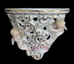 ARTE SACRA, Peanha de parede em porcelana esmaltada na tonalidade branca, ricamente decorada com elementos vegetalistas, vazados e querubins em relevo e policromia, realces em ouro, finalizada por volutas, 1 asa colada. Med.: 13,5x16,5cm.