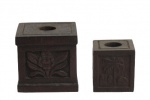 IVOTICI, Dois castiçais confeccionados em madeira, formato quadrangular, 1 com entalhes de flores e folhagens e 1 com entalhes de indios e palmeiras. Med.: 5,5x5,5cm / 4x4cm.