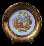 DP 541, Prato decorativo em porcelana esmaltada, decorada em relevo, borda em vasta douração, parte central com cena maternal em rica policromia. Med.: 26cm.
