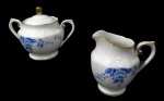 NORITAKE, Açucareiro e leiteira, confeccionadas em porcelana esmaltada na tonalidade branca, arabescos realçados em relevo, pintura de flores e folhagens na tonalidade azul e bordas filetadas a ouro. Med.: Açucareiro: 10x13x7cm / Leiteira: 10x10x5,5cm.