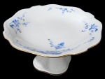 NORITAKE, Fruteira de pé, confeccionada em porcelana esmaltada na tonalidade branca, arabescos realçados em relevo, pintura de flores e folhagens na tonalidade azul e borda filetadas a ouro. Med.: 12x22x22cm.