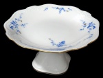 NORITAKE, Fruteira de pé, confeccionada em porcelana esmaltada na tonalidade branca, arabescos realçados em relevo, pintura de flores e folhagens na tonalidade azul e borda filetadas a ouro. Med.: 12x22x22cm.