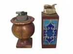 DIVERSOS, Dois isqueiros, sendo: A) EVANS, EUA, Isqueiro de mesa, confeccionado em metal dourado, suporte em madeira, placa decorativa em cloisone. Não testado e sem garantia de funcionamento. Med.: 14,5cm, e B) 1 isqueiro de mesa, confeccionado em metal dourado, suporte em madeira representando vaso, decorado em gomos, apoiado sobre base quadrada. Med.: 12cm. Ambos apresentam sinais de oxidação.