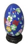 OBJETOS DECORATIVOS, Ovo confeccionado em cloisone, esmaltado na tonalidade azul, ricamente decorado por flores e folhagens em policromia, apoiado sobre peanha em madeira. Med.: 16cm.