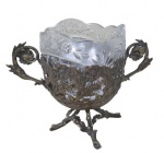 METAL PRATEADO, Uveira de pé, confeccionada em metal espessurado a prata, ricamente cinzelada por flores e folhagens com vazados, pés no feitio de tronco, recipiente adaptado em cristal translucido, lapidado por sulcos, flores e folhagens, borda em satine e fundo estrelado. Necessita banho. Med.: 14x18x10cm.