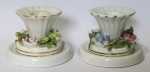 P. II, Rio de Janeiro, Par de castiçais para 1 vela, confeccionados em porcelana esmaltada na tonalidade branca, decorado em gomos, flores e folhagens em relevo e policromia, base circular escalonada. Ambos possuem fio de cabelo na bobeche, 1 possui craquele e ínfimos bicados nas flores. Med.: 7x6,5cm.
