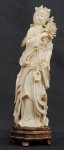 KUAN YIN - ELABORADA ESCULTURA EM MARFIM REPRESENTANDO FIGURA DE KUAN YIN A DEUSA DA MISERICÓRDIA.  SEGURA NA MÃO ESQUERDA GRANDE RAMO COM PEÔNIA E NA MÃO DIREITA UM ABANADOR. ASSENTE SOBRE BASE EM MADEIRA. CHINA, SEC. XIX. ). 24 CM DE ALTURA. MINIMOS BICADOS