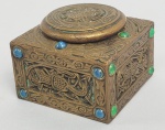 TIFFANY STUDIOS MODELO 1699 - "NINTH CENTURY" (SÉCULO IX)  TINTEIRO EM BRONZE COM FEITIO QUADRADO ESTILO E ÉPOCA ART DECÔ. O MODELO "NINTH CENTURY"  É DECORADO COM MOTIVOS CELTAS MEDIEVAIS ZOOMMORFOS, INCRUSTADOS COM CABOCHONS  DE VIDROS COLORIDOS FAVRILE, SIMULANDO PEDRAS PRECIOSAS. INTERIOR COM FRASCO EM VIDRO ARTÍSTICO. SOB A BASE MARCA DA MANUFATURA TIFFANY STUDIOS NEW YORK 1699. NOVA YORK, CIRCA DE 1912. 7 X 6 CM  . Nota: Mestre em quase todos os meios aos quais recorreu, Louis Comfort Tiffany esteve à frente de várias empresas diferentes que levam seu nome ao longo de sua longa carreira de cinco décadas nas artes plásticas e decorativas. Os vários empreendimentos artísticos de Tiffany tomaram forma no final da década de 1870, quando ele formou a Louis C. Tiffany & Company, projetando interiores artísticos e móveis sob medida no gosto estético para alguns dos membros mais ricos e intrigantes da sociedade da Era Dourada de Nova York, incluindo Henry Osborne. Havemeyer, George Kemp, Ogden Goelet, Mark Twain e outros luminares - Tiffany até ganhou a encomenda do presidente Chester A. Arthur para decorar o interior da Casa Branca em 1882. Embora não esteja claro quando ele começou a experimentar o meio, o vidro começou a aparecer em seus inovadores mobiliários artísticos nesses interiores rarefeitos. Ele incorporou painéis de vidro com chumbo em móveis ricamente esculpidos bem como painéis arquitetônicos maiores com motivos estampados, como em sua encomenda de 1880 com Candace Wheeler para o Union League Club e um painel monumental para a Casa Branca, agora perdido. Tiffany começou a desenhar composições mais pictóricas na década de 1880, frequentemente retratando flora e fauna, e mais tarde paisagens, em vidro com chumbo.  O boca a boca levou ao aumento da demanda e à criação da Tiffany Glass Company e, mais tarde, da Tiffany Glass and Decorating Company. Embora esses primeiros trabalhos tenham sido fabricados com painéis de vidro plano adquiridos, os fabricantes comerciais de vidro não conseguiram produzir os efeitos texturais e visuais que Louis Comfort Tiffany desejava. Para alcançar sua visão artística, Tiffany vasculhou o mundo em busca dos melhores cientistas, químicos e sopradores de vidro e, no início da década de 1890, reuniu-os para formar a Stourbridge Glass Company em Corona, Queens, o início de um grande complexo de fabricação personalizado que iria está no centro de seu império de design. Uma série de exibições de sucesso em Feiras Mundiais - nas quais Tiffany e seus designers arrecadaram Grandes Prêmios quase todos os anos em que expuseram - apenas permitiu que os negócios da Tiffany florescessem ainda mais e, no final de 1902, ele fundou a empresa pela qual é mais conhecido hoje: Estúdios Tiffany. Tiffany e os numerosos designers e artesãos experientes que ele empregou na Tiffany Studios (e mais tarde na Louis C. Tiffany Furnaces) projetaram e produziram alguns dos exemplos mais icônicos do design americano na virada do século XX. Além das populares lâmpadas Tiffany de vidro com chumbo, como a Dragonfly e a Wisteria , a empresa produziu milhares de magistrais vidros Tiffany, bem como os mosaicos de vidro arquitetônicos Favrile instalados em edifícios públicos e privados nos Estados Unidos. Embora menos conhecidos, Tiffany e seus artesãos foram responsáveis por uma série de inovações técnicas tanto em vidro soprado quanto em esmaltagem, muitas vezes resultados de experimentos dispendiosos realizados para alcançar o efeito visual desejado por Tiffany. 