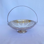 REGIA - Cesta redonda com bordas recortadas em metal espessurado a prata com detalhes cinzelados de arabescos e volutas e alça articulada. Med.: 8cm de altura e 24,5cm de diâmetro. Apresenta desgastes.