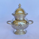 Antigo açucareiro em metal espessurado a prata, decorado com volutas, tampa com pegador em pinha e duas alças, med. 15cm de altura