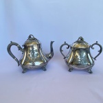 INICIO SEC. XX - Bule e Açucareiro em metal espessurado a prata com decorações de folhas de pareiras cinzeladas e pegadores em formato de fruta sobre folha e alças em volutas, med. 18cm e 17cm de altura. Apresenta desgaste do tempo, pegador da tampa do bule faltando a porca interna.