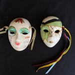 Par máscaras sendo uma em porcelana e uma em cerâmica, pintadas a mão com detalhes em gliter, med. 8,5X12cm e 9X13cm. Manchas nos detalhes da pintura.