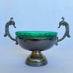 MAJEDI - Pequeno centro de mesa redondo em metal espessurado a prata, com bowl em vidro verde no centro e 2 pegas em formato de cavalo marinho. Medida 15cm de altura, 14cm. de diâmetro e 22cm de largura. Bowl com trinca