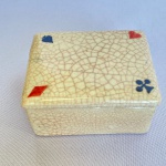 Caixa com tampa em porcelana craquelada com tampa decorada com símbolos de naipes. Med.: 4,5cm de altura e 6cm de largura e 8cm de comprimento. Apresenta bicado na base