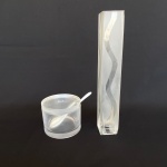 Lote com 2 (duas) peças sendo: Um açucareiro acrílico grosso com colher de porcelana branca (com bicado); E um vaso para flores quadrado de vidro (possui bicados). Med.: 7cm de altura e 9cm de diâmetro; e 25cm de altura e 4cm de largura.