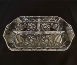 Petisqueira retangular com alças laterais e três divisões em cristal translúcido prensado em formas geomêtricas e lapidações de estrelas e filetos. Med.: 3,5cm de altura e 30cm de comprimento.