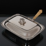 <b>Panela "réchaud"</b> conhecida como "cheese dish" em prata inglesa do período George III. Apresenta grande brasão armorial lavrado na face anterior da tampa. Pega modelada em coroa sobre almofada. Borda em godrões intercalados por volutas e "coquilles". Alça destacável em marfim belamente entalhado. Contraste da cidade de Londres de 1817 e marca ilegível de prateiro. Compr. 28 cm; Prof. 21 cm. Peso 1400 gramas.