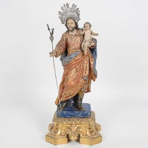 Bela imagem portuguesa de <b>São José de Botas com o Menino Deus</b> em madeira. Vestes e manto em pregueados, ricamente policromados e realçados a ouro com volutas e estrelinhas. Coturnos na cor preta. O Menino Jesus encontra-se apoiado em seu braço esquerdo e um cajado com lírios à mão direita. Base entalhada com elementos barrocos a ouro. Resplendor e cajado em prata. Séc. XVIII. Os braços do menino colados. Alt. sem o resplendor 37,5 cm