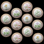 <B>Conjunto de 12 mini pratos para manteiga </b>em porcelana dinamarquesa da importante manufatura Royal Copenhagen, modelo <b>"Flora Danica"</b>. Cada peça, inteiramente pintada à mão em belíssima policromia com suas designações botânicas e numeração no verso. Apresentam bordas em mínimos picotados cobertos a folha de ouro. Diâm. 7,5 cm