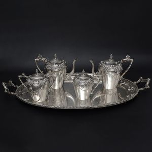 <B>Serviço p/ chá e café</b> em prata portuguesa com parte superior das peças decoradas com estilizações e arcadas de linhas Art Nouveau. Bordas circundadas por cordões e pegas das tampas modeladas em pinhas. Centro da bandeja monogramado. Isoladores em marfim. Constando de: bandeja, 2 bules, leiteira e açucareiro. Contraste Javali da cidade do Porto usado de 1886 a 1938. Compr. da bandeja 81 cm. Alt. do bule maior 20 cm. Peso 6200 gramas.