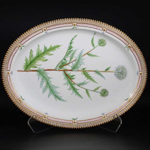 Grande <b>travessa oval </b>em porcelana dinamarquesa da importante manufatura Royal Copenhagen, modelo "Flora Danica". Inteiramente pintada à mão em belíssima policromia com suas designações botânicas e numeração no verso. Apresenta borda em mínimos picotados cobertos a folha de ouro. Med. 47 x 36,5 cm