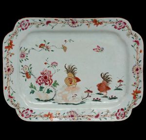 <B>Travessa</b> de forma retangular com cantos arredondados em porcelana Companhia das Índias pertencente ao "Serviço dos Galos de D. João VI". Ao centro, em policromia, duas figuras de galos e borboleta cercados de flores. Borda com ramagens também floridas entrelaçadas em galhos de bambu. Séc. XVIII. Apresenta armação metálica para parede. Med. 36 x 26,5 cm